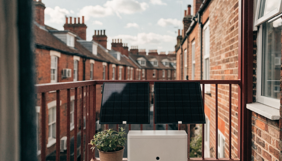Plug-in solar panels on the railing of a UK flat's balcony with a compact plug-in battery beside them, British brick terraces and chimney pots in the background